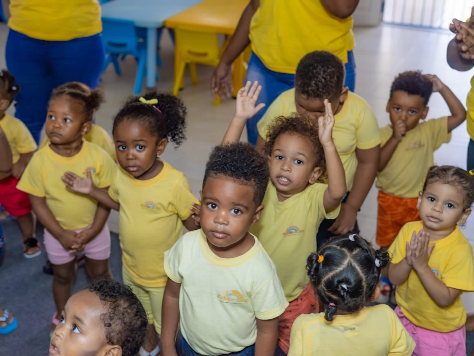 Toddlers in yellow KinderLand shirts looking up at the camera, one with a hand raised